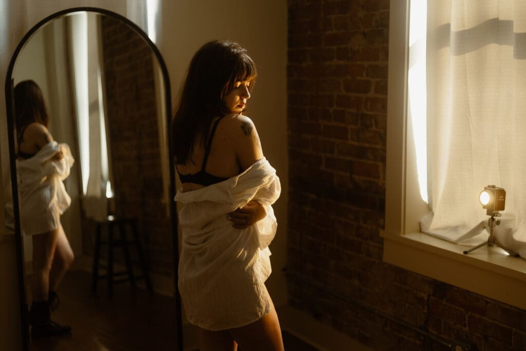 woman wrapped in white flowy shirt in front of a mirror during golden hour boudoir session at the lightfoot studio in knoxville