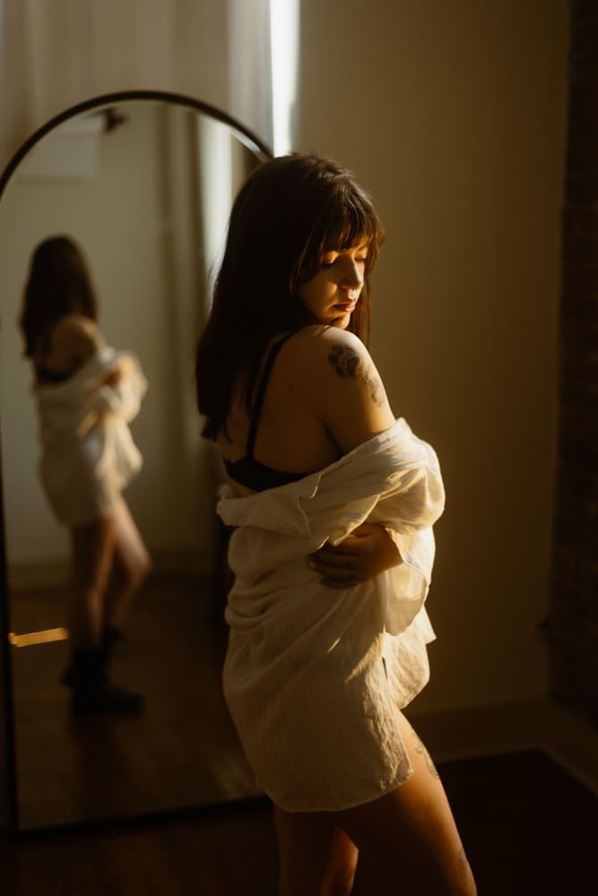 woman wrapped in white flowy shirt in front of a mirror during golden hour boudoir session at the lightfoot studio in knoxville